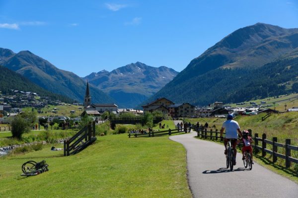 Due persone in bicicletta lungo un sentiero montano con un villaggio sullo sfondo.