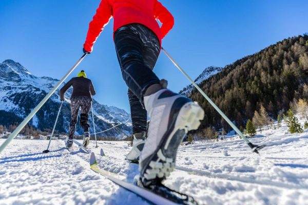 Due persone fanno sci di fondo su un sentiero innevato con montagne sullo sfondo.