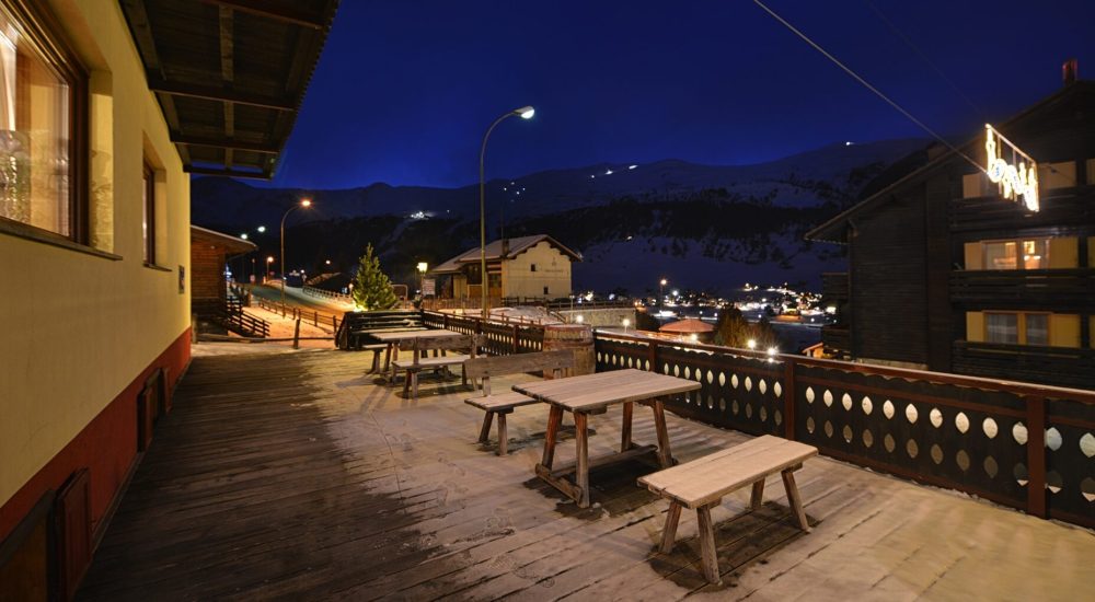 Terrazza esterna di una baita di montagna vista di notte con tavoli in legno e paesaggio innevato.
