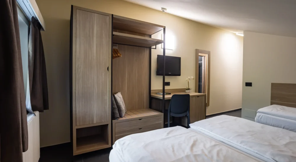 Modernes Hotelzimmer mit zwei Einzelbetten, einem Schreibtisch, einem offenen Kleiderschrank und einem an der Wand montierten Fernseher.